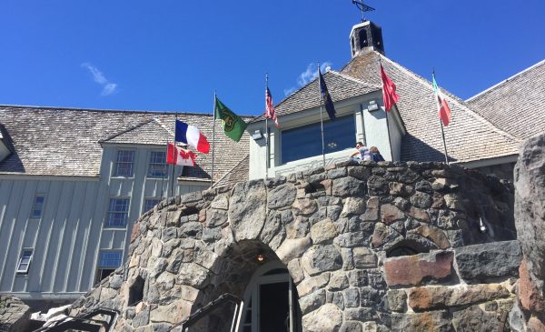 Seven flags stick up from the edge of the second floor deck of Timberline Lodge. The main entrance of Timberline is made of local stone and two people are sitting on the deck above the main entrance.