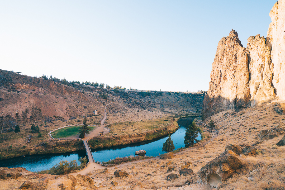 Blue sky above a wooden footbridge crossing a winding blue river. Surrounding the river are dried brown grasses, dotted with dark brown rocks. Rising from the ground and jutting toward the sky are magnificent red-brown rocks used by rock climbers. 